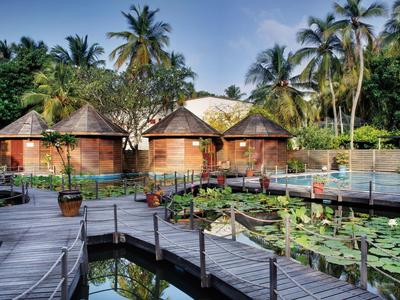 Drei runde Holzhäuser mit Strohdächern, umgeben von Palmen und Holzstegen über einem Wasserteich.
