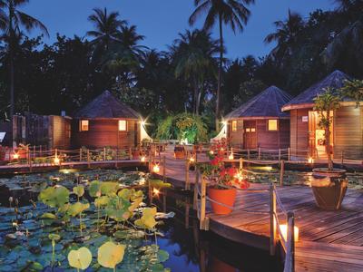 Holzhäuser am Wasser mit Seerosen, Palmen und warmem Licht in tropischer Nacht.