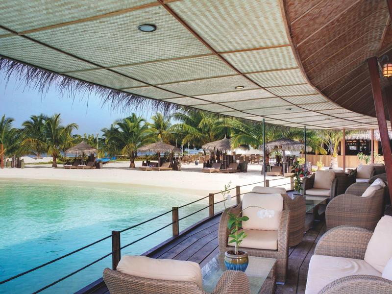 Überdachte Lounge mit weißen Sofas und Blick auf Pool und Palmen am Strand.