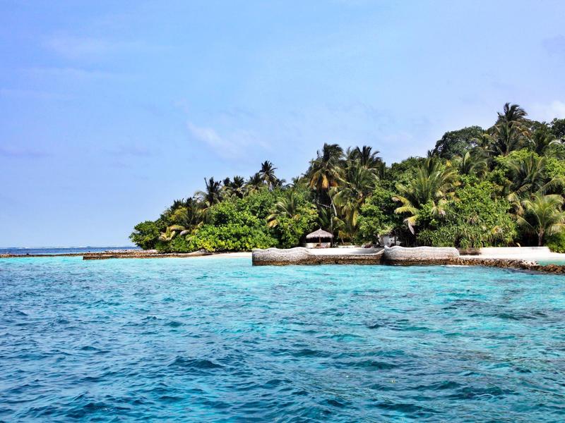 Klares blaues Meer vor tropischer Insel mit dichtem Grün und Sandstrand unter blauem Himmel.