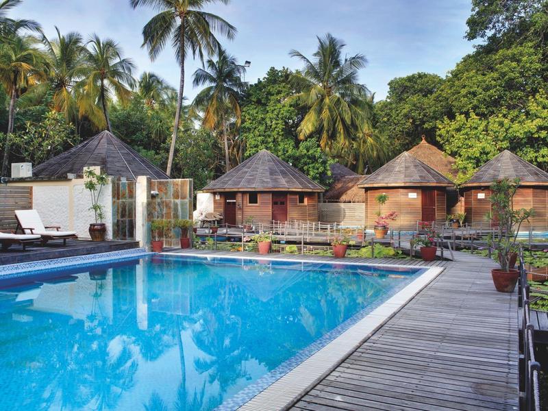 Schwimmbad mit klar blauem Wasser, umgeben von Holzstegen und tropischen Hütten mit Palmen im Hintergrund.