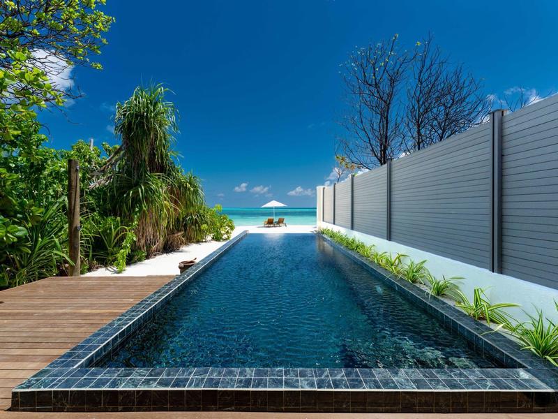 Langgezogener schwarzer Pool mit Holzsteg, grünen Pflanzen und blauem Himmel am Meer.