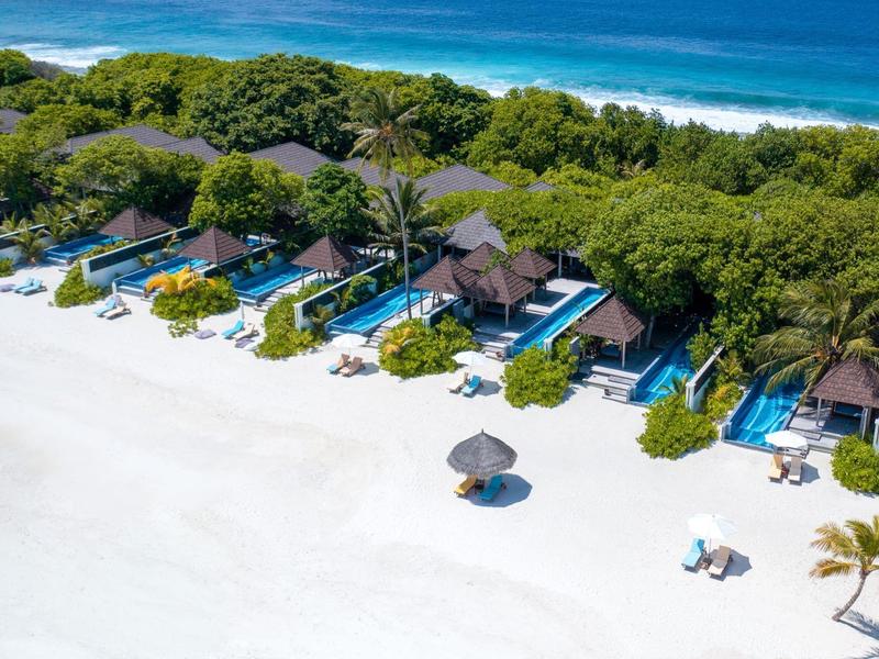 Strandbungalows mit grünen Dächern an weißem Sandstrand, hinter ihnen dichter grüner Wald und blaues Meer.
