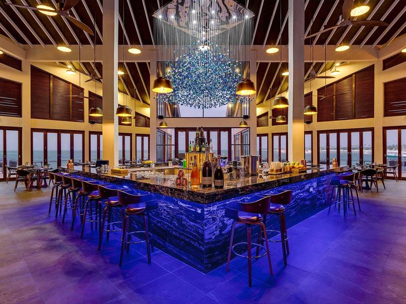 Moderner Barbereich mit blau beleuchtetem Tresen, Holzstühlen und großer Glaslampe an der Decke.