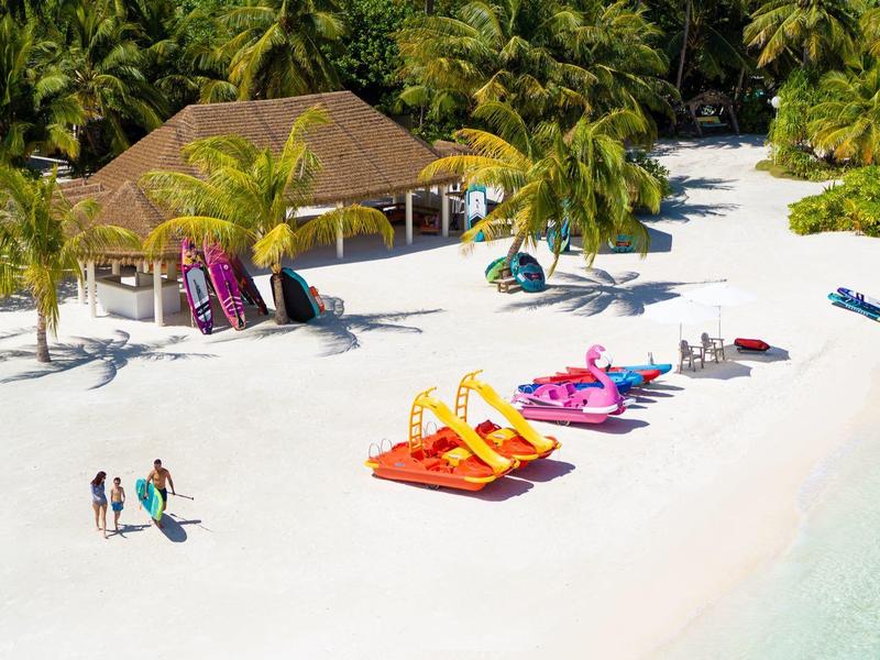 Strand mit weißem Sand, bunten Kajaks, Palmen und einem kleinen Gebäude im Hintergrund.