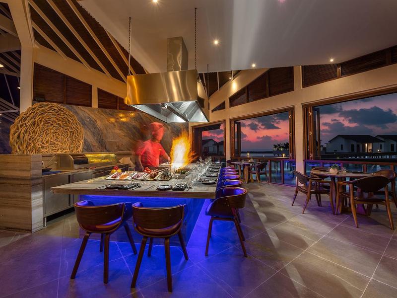 Modernes Restaurant mit offenem Kochbereich, blauen Lichtern und Panoramafenstern bei Sonnenuntergang.