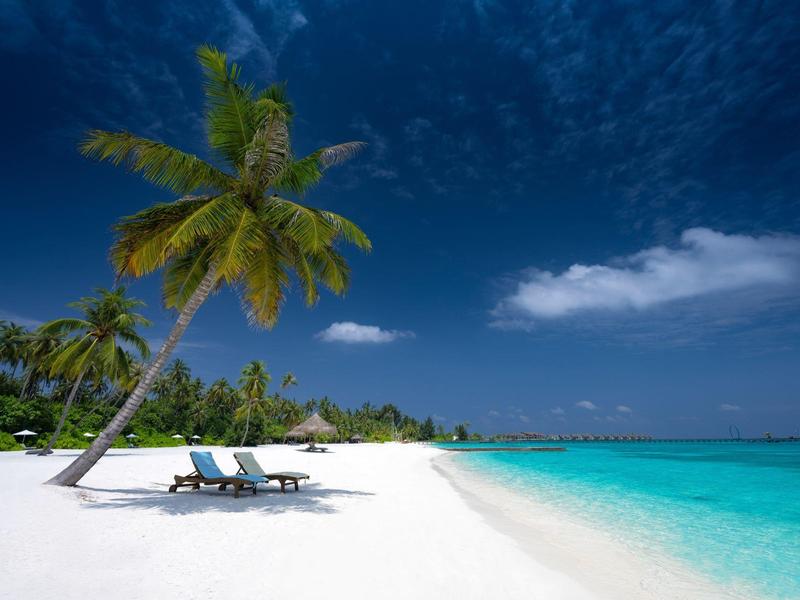 Strand mit weißem Sand, türkisfarbenem Meer, bewölktem Himmel und Palme mit zwei Liegestühlen.