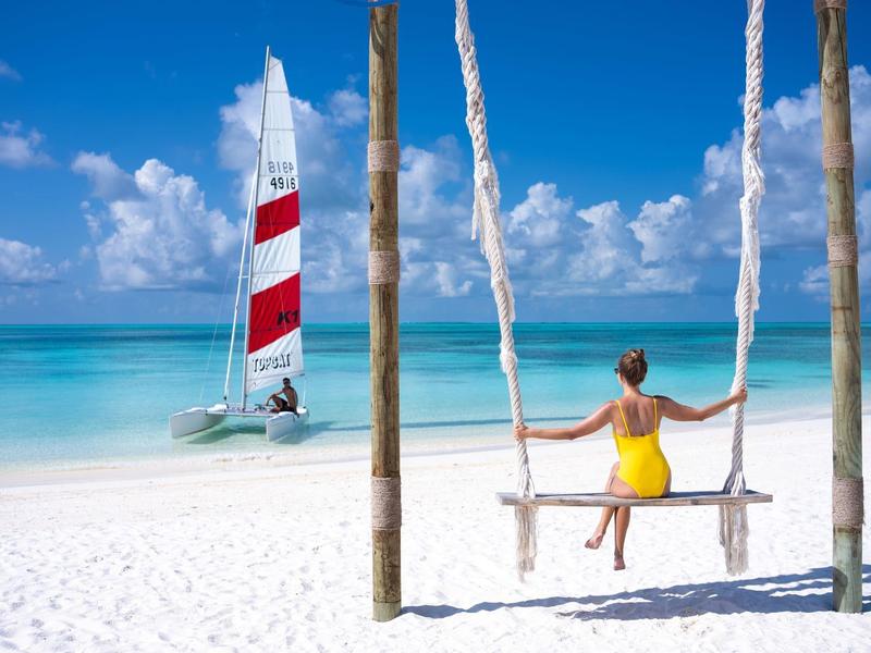 Frau in gelbem Kleid auf Holzschaukel am weißen Strand mit blauem Meer und rotem Segelboot.
