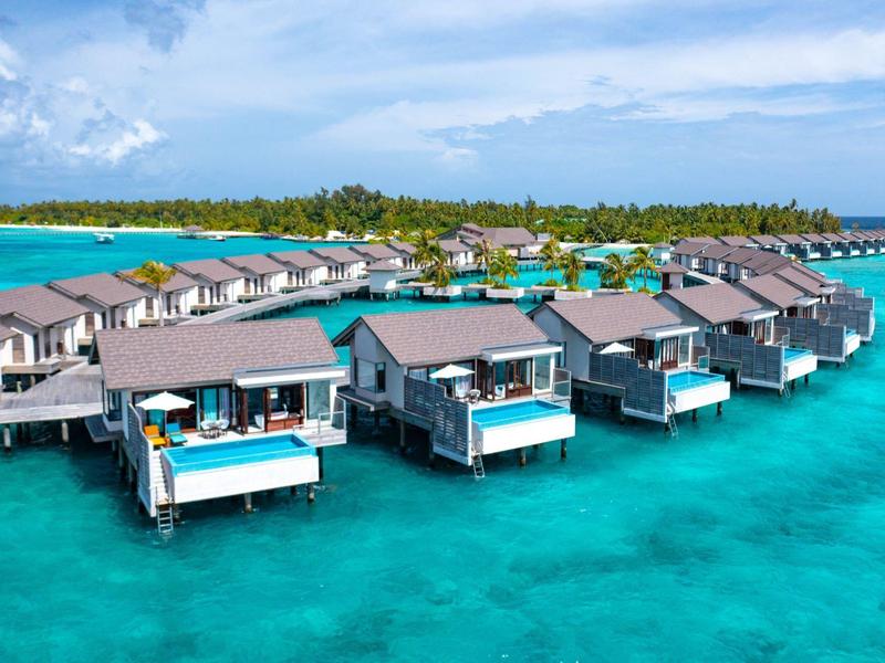 Reihen von Wasservillen auf Pfählen mit Terrassen über klarem türkisblauem Meer vor Inselkulisse.