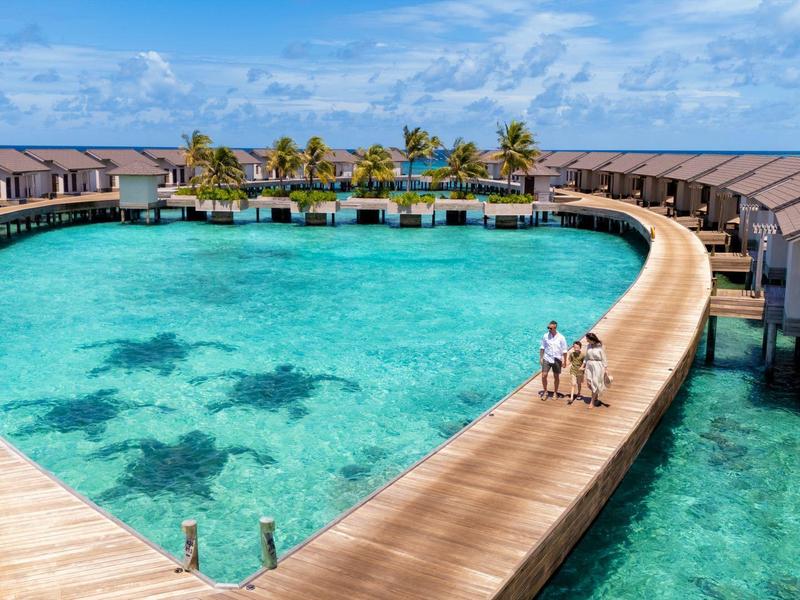 Herzförmige Überwasserbungalows mit Holzsteg, klar blaues Wasser und Palmen im Hintergrund.