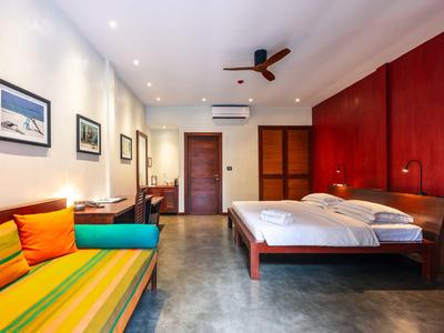 Helles Schlafzimmer mit Doppelbett, bunter Sofa, Ventilator an der Decke und Holzmöbeln an der Wand.