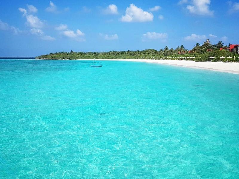 Türkisfarbenes klares Wasser und weißer Sandstrand unter blauem Himmel mit vereinzelten Wolken.