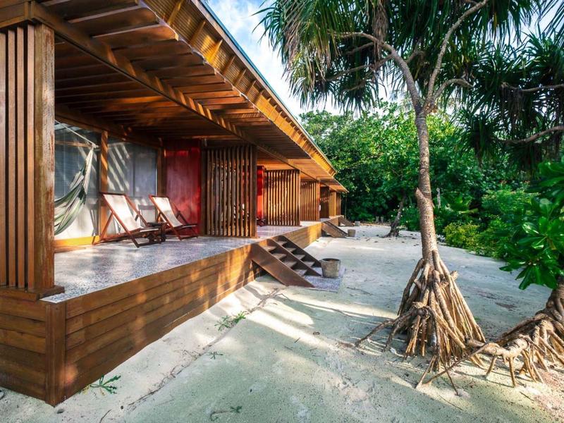 Holzhaus mit Veranda am Sandstrand, umgeben von grünen Palmen und tropischem Bewuchs.