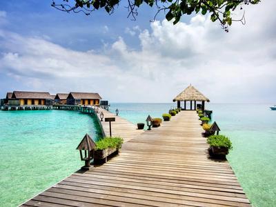 Pontile di legno che conduce a un piccolo padiglione su acque tropicali limpide con bungalow sullo sfondo.