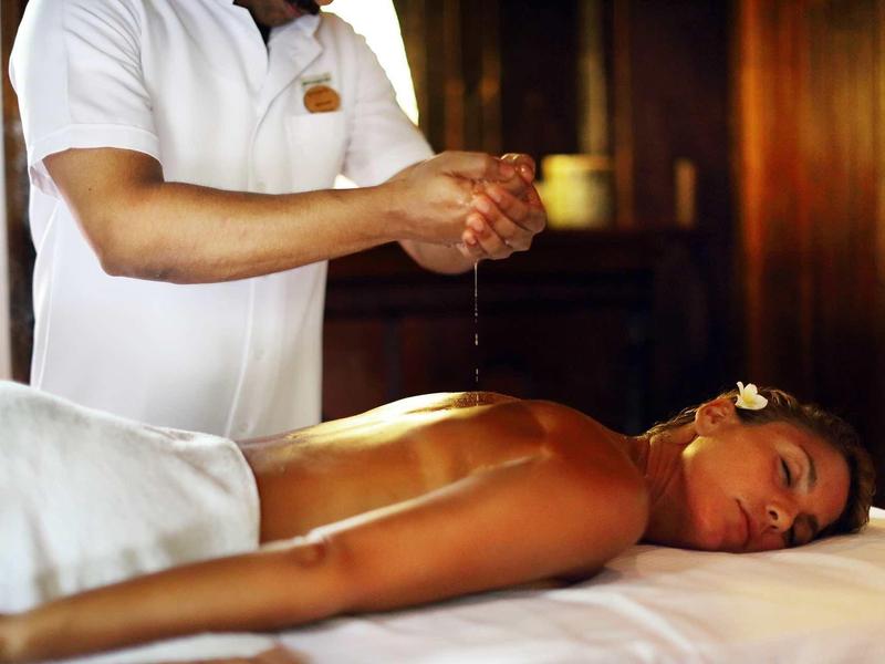 Frau liegt entspannt auf Massagetisch, während Therapeut Öl auf ihre Hände gibt, warme Holzumgebung.