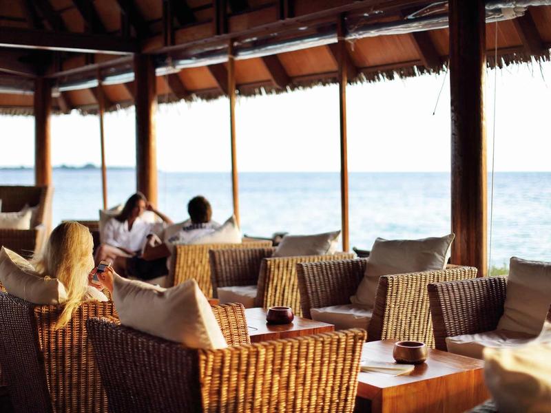 Offenes Strandcafé mit Rattansesseln, Holztischen und Blick aufs blaue Meer am hellen Tag.