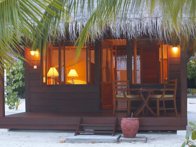 Kleine Holzhütte mit Strohdach, beleuchteten Fenstern und Veranda mit Tisch und Stühlen.