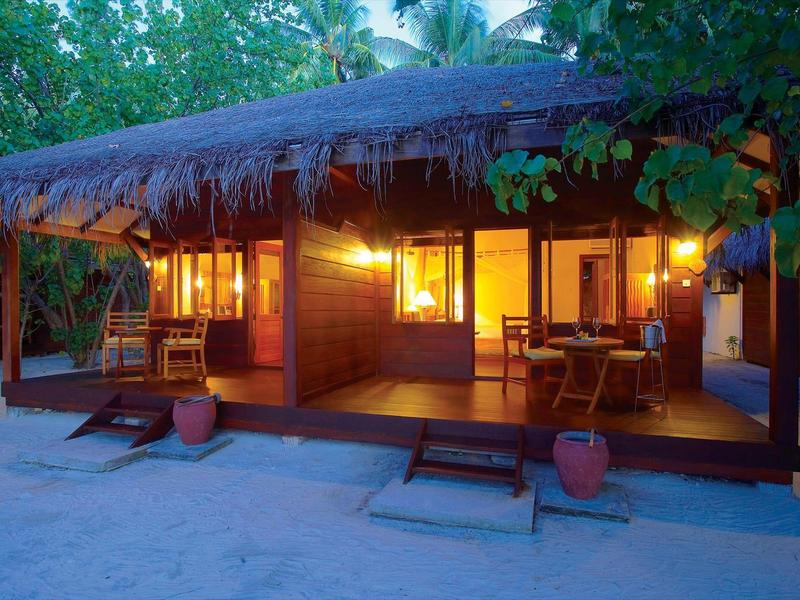 Holzhütte mit Strohdach, warm beleuchtet, umgeben von Bäumen und Sandboden in der Dämmerung.