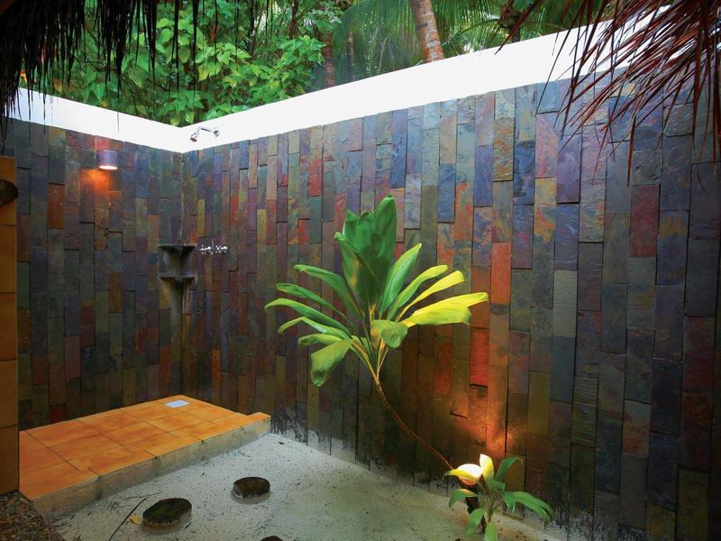 Offene Dusche mit Natursteinwänden, grünem Busch und dekorativem Licht am Boden.