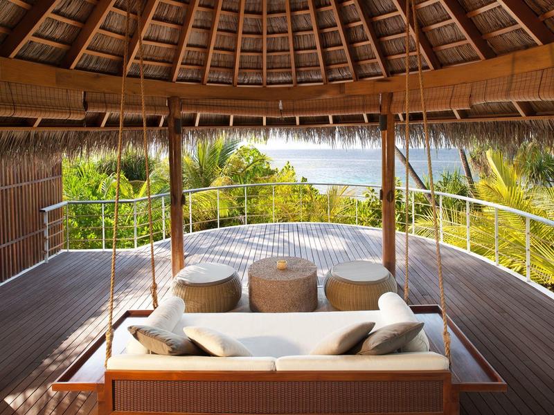 Hängende Lounge-Sitzbank unter einem strohgedeckten Dach mit Blick auf Palmen und Meer.