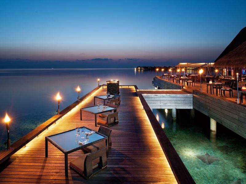 Abendliche Terrasse eines Hotels am Meer mit beleuchteten Tischen und ruhiger Wasserfläche.