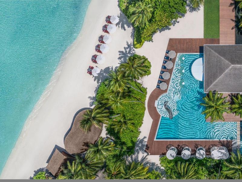 Luftaufnahme eines tropischen Resorts mit Pool, Palmen und weißem Sandstrand am türkisfarbenen Meer.