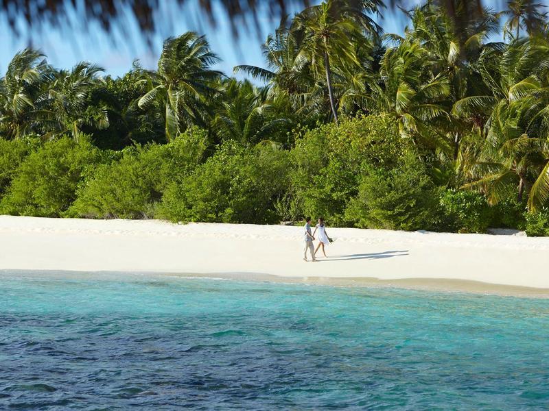 Zwei Personen laufen am weißen Sandstrand entlang, umgeben von tropischen Palmen und klarem blauem Wasser.
