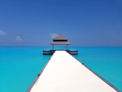 Weißer Steg führt zu einer kleinen Hütte über türkisfarbenem Meer unter blauem Himmel.