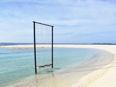 Schaukel hängt über klarem, flachem Wasser an einem weißen Sandstrand unter blauem Himmel.