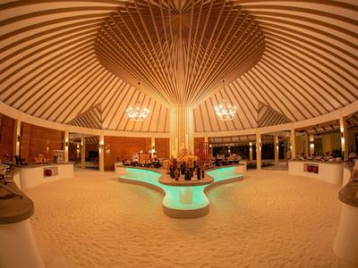 Innenraum einer kreisförmigen, hölzernen Hotel-Lobby mit zentralem, beleuchtetem Brunnen und Sandboden