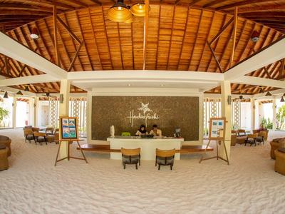 Geräumige Hotellobby mit weißem Sandboden, Holzdach und komfortablen Sitzbereichen.