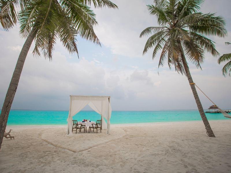 Weißer Sandstrand mit Palmen, einer großzügigen überdachten Sitzgelegenheit und türkisfarbenem Meer im Hintergrund.