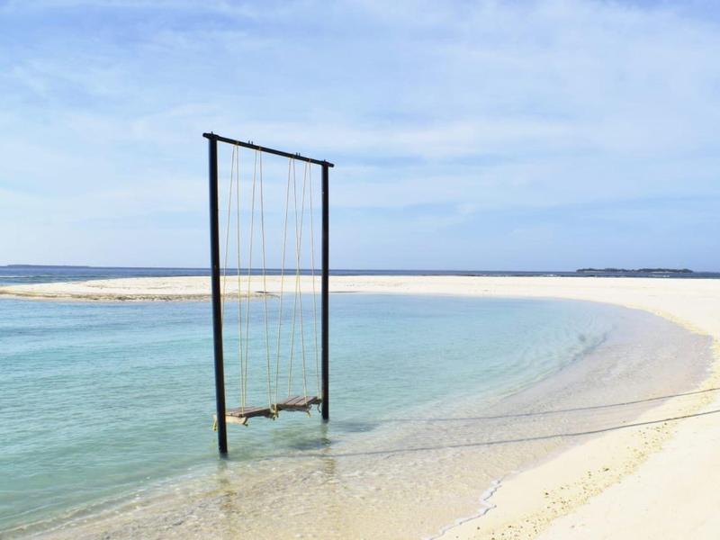 Schaukel hängt über klarem, flachem Wasser an einem weißen Sandstrand unter blauem Himmel.
