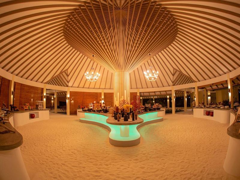 Innenraum einer kreisförmigen, hölzernen Hotel-Lobby mit zentralem, beleuchtetem Brunnen und Sandboden