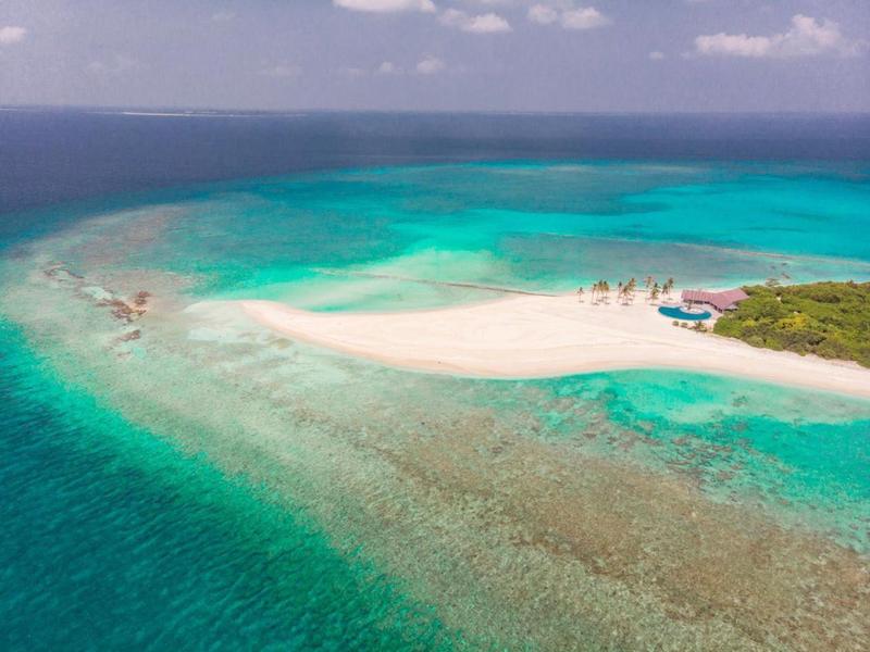 Kleine Sandinsel umgeben von türkisfarbenem, klarem Wasser und grünem Bewuchs am Rand.