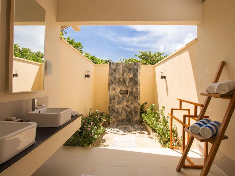 Offenes Badezimmer mit Dusche, Waschbecken und Handtüchern, umgeben von Wänden und Pflanzen.