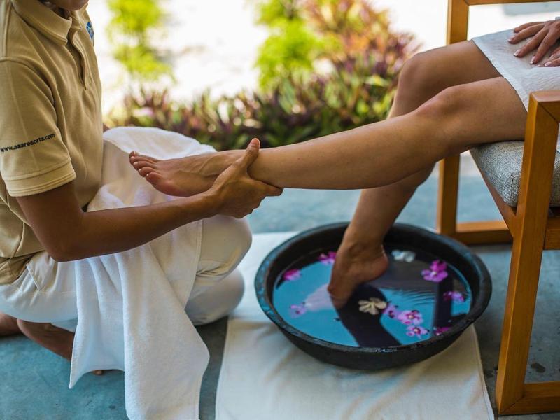 Person massiert Fuß einer anderen, die in Schale mit Wasser und Blüten sitzt, helle Kleidung, helles Umfeld.