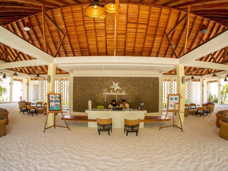 Geräumige Hotellobby mit weißem Sandboden, Holzdach und komfortablen Sitzbereichen.