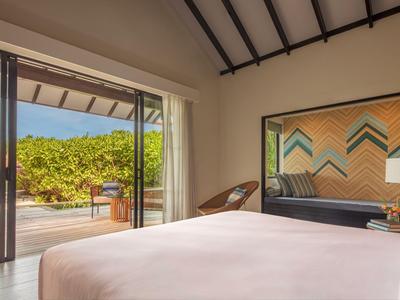 Jasny pokój hotelowy z dużym łóżkiem, krzesłem i wyjściem na taras z widokiem na zielone rośliny.