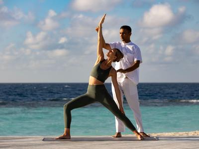 Kobieta ćwiczy jogę na plaży, prowadzona przez mężczyznę, z morzem w tle.