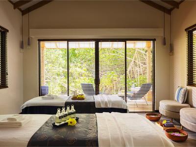 Jasny pokój hotelowy z dwoma łóżkami, sofą i widokiem na zielony ogród przez duże okna.