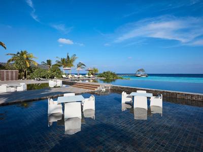 Piscina a sfioro con sedute bianche e vista sul mare e bungalow sulla spiaggia sotto un cielo limpido.