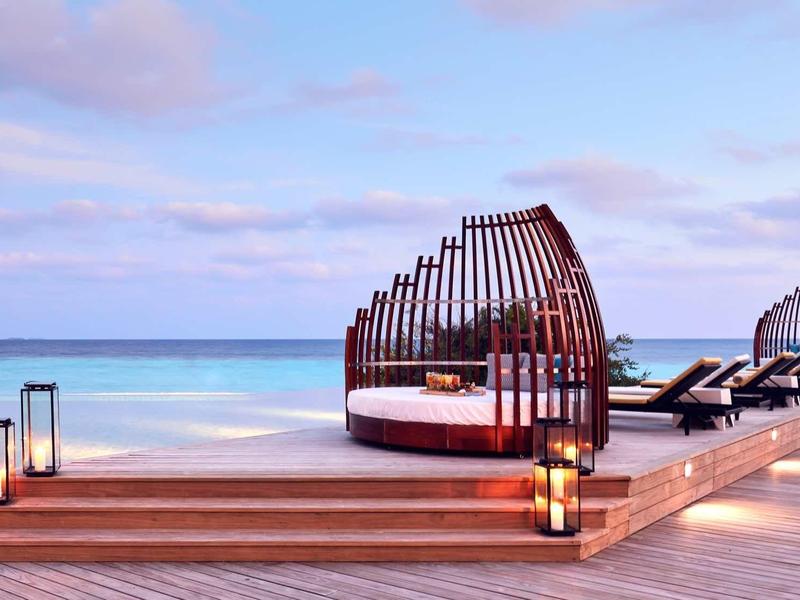 Eleganti letti da spiaggia con archi di legno e lanterne su un pontile con vista mare al tramonto.
