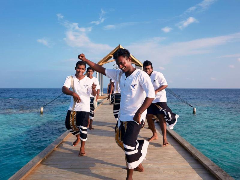 Tre uomini in abiti tradizionali danzano su un molo sul mare sotto un cielo azzurro.