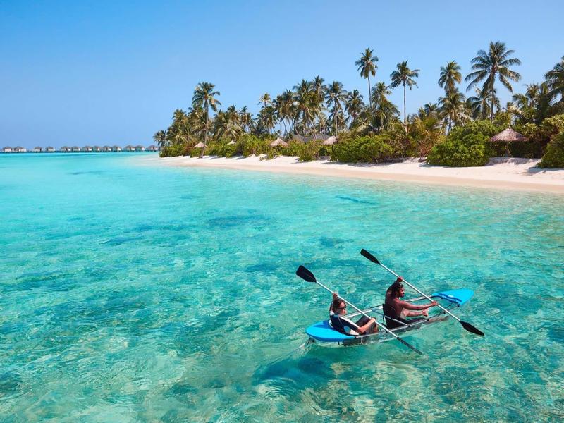 Due persone in kayak su acqua turchese limpida vicino a una spiaggia di sabbia bianca con palme.