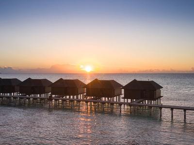 Sonnenuntergang über Bungalows auf Stelzen im Meer, verbunden durch einen Steg.