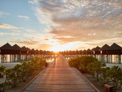 Ein Steg führt bei Sonnenuntergang zu Überwasserbungalows in tropischer Umgebung.