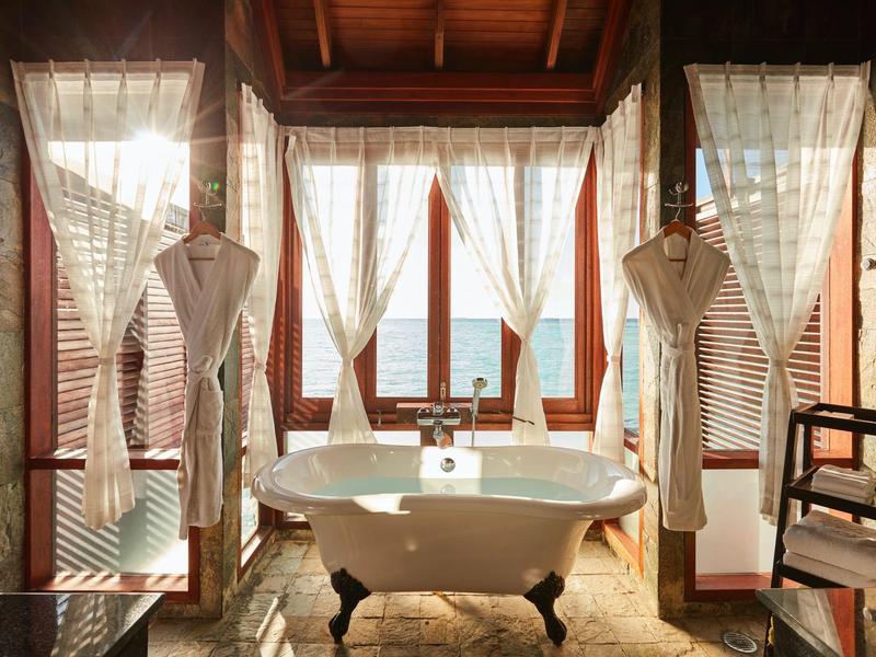 Vasca da bagno autoportante davanti a grandi finestre con vista sull'acqua, accappatoi appesi.
