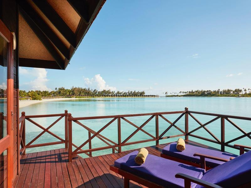Veranda in legno con lettini viola di fronte a acqua calma e palme.