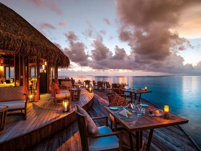 Holzdeck mit Essbereich und Sitzgelegenheiten neben ruhigem Meer bei Sonnenuntergang.
